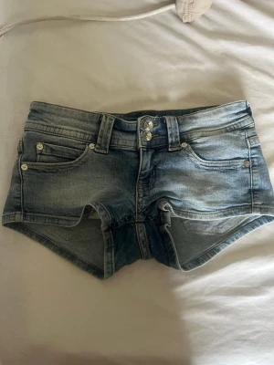 Blå denimshorts med knappar - Säljer ett par blå denimshorts med låg midja och dubbla knappar framtill. Shortsen har klassiska fem fickor, bälteshällor och snygga detaljer på bakfickorna med knappar. Perfekta för varma dagar och en avslappnad stil.