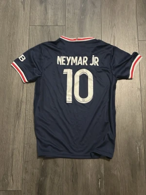 PSG Neymar Jr matchtröja blå - Säljer en mörkblå Paris Saint-Germain matchtröja med Neymar Jr och nummer 10 på ryggen. Tröjan har vita och röda detaljer vid krage och ärmslut samt PSG-logga och sponsortryck på bröstet. Perfekt för dig som älskar fotboll och vill stötta PSG.
