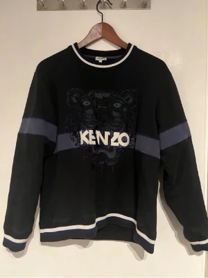 Svart Kenzo sweatshirt med tiger - Svart sweatshirt från Kenzo med ikonisk tigerbrodyr och stort Kenzo-tryck på bröstet. Tröjan har blå och vita detaljer på ärmar och nederkant samt ribbade muddar. Perfekt för dig som vill ha en statement piece i garderoben.