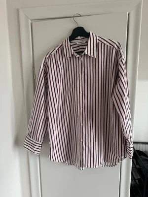 Randig skjorta från Kappahl - Säljer en vit skjorta med vinröda vertikala ränder från Kappahl. Skjortan har klassisk krage, långa ärmar och knäppning framtill. Perfekt för dig som gillar en stilren och trendig look.
