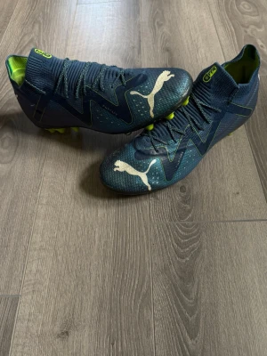 Blåa Puma fotbollsskor med neon - Säljer ett par blåa Puma fotbollsskor med vita loggor och neonfärgade detaljer. Skorna har stickad strumpliknande krage, snörning och mönstrad ovandel för extra grepp. Sulan har neonfärgade dobbar som passar perfekt för spel på konstgräs.