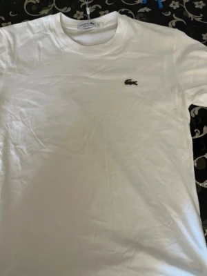 Vit t-shirt från Lacoste - Klassisk vit t-shirt från Lacoste med rund hals och den ikoniska broderade krokodilloggan på bröstet. Tillverkad i mjuk bomull för en skön känsla och enkel stil. Perfekt basplagg för alla tillfällen.