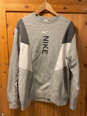 Grå sweatshirt från Nike - Snygg grå sweatshirt från Nike med vita och mörkgrå paneler på ärmarna. Stor Nike-logga och text framtill. Tröjan har rund halsringning, ribbade muddar och är tillverkad i mjuk bomullsblandning. Perfekt för en sportig och avslappnad stil.