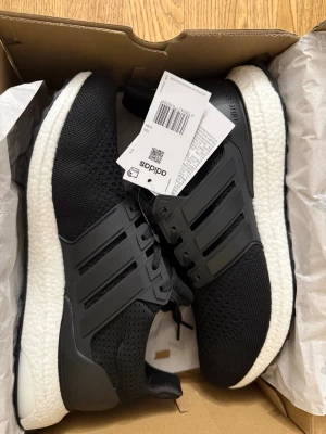Svarta Adidas Ultraboost sneakers - Snygga svarta Adidas Ultraboost sneakers med stickad ovandel och klassiska tre ränder på sidan. Skorna har en vit, responsiv Boost-sula och svarta detaljer i plast runt snörningen. Perfekta för dig som gillar sportig och modern stil.