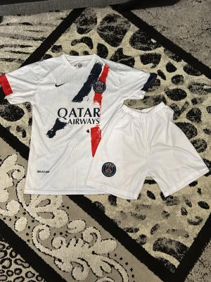 PSG fotbollskit Kvaratskhelia #7 - Snyggt PSG matchställ med vit tröja och shorts. Tröjan har röda och blå detaljer, Qatar Airways-logga och PSG-märke på bröstet. Nummer 7 och Kvaratskhelia på ryggen. Tillverkad i lätt och ventilerande material, perfekt för fotbollsträning eller match.