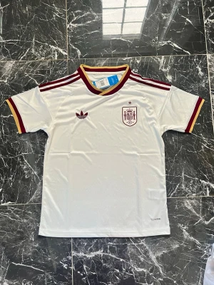 Vit Spanien fotbollströja Adidas - Säljer en vit Spanien fotbollströja från Adidas med vinröda och gula detaljer. Tröjan har korta ärmar, rund halsringning och klassiska Adidas-ränder på axlarna. Tillverkad i ett lätt och ventilerande material, perfekt för träning eller match.