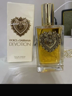 Dolce & Gabbana Devotion EdP - Lyxig parfym från Dolce & Gabbana, Devotion Eau de Parfum. Flaskan är fyrkantig i genomskinligt glas med guldfärgat lock och en detaljerad hjärtformad metallprydnad framtill. Doften har en varm, gyllene ton och förpackningen är elegant med matchande design.