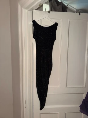 Svart glittrig långklänning - Snygg svart långklänning med glittrigt tyg och asymmetrisk nederkant. Klänningen har breda axelband, v-ringad rygg och en cool cut-out detalj i ryggen. Perfekt för dig som vill sticka ut på festen!