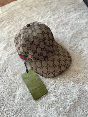 GG keps - Säljer en beige keps från GG med klassiskt GG-monogram över hela kepsen. Kepsen har en böjd skärm och justerbar rem bak. På sidan finns den ikoniska gröna och röda randen. Materialet är canvas med läderdetaljer.