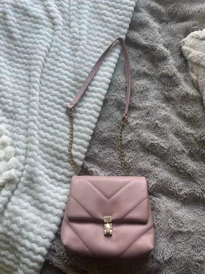 Ljusrosa quiltad handväska - Söt ljusrosa handväska i quiltad design med guldfärgad kedja och spänne. Väskan har en mjuk, matt finish och ett elegant geometriskt mönster på framsidan. Perfekt för dig som gillar stilrena accessoarer med en feminin touch.
