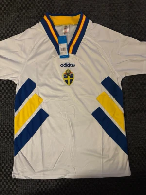 Svensk landslagströja Adidas vit M - Vit svensk landslagströja från Adidas med blå och gula detaljer samt SVFF-emblem på bröstet. Tröjan har V-ringad krage med blå, gula och röda ränder. Tillverkad i lätt och ventilerande material, perfekt för fotboll eller träning.