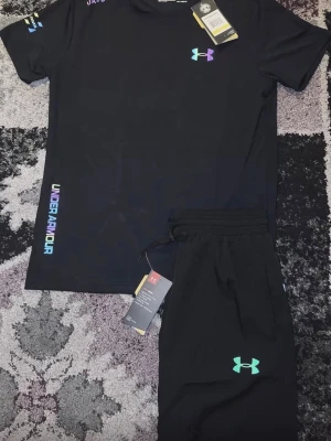 Svart träningsset från Under Armour - Snyggt svart träningsset från Under Armour med t-shirt och shorts. Setet har färgglada loggor och textdetaljer i neonfärger. Tillverkat i ett lätt och snabbtorkande material som passar perfekt till träning eller sport. 