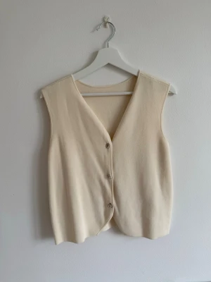 Beige stickad väst med knappar - En stilren beige stickad väst med v-ringning och tre knappar framtill. Västen har en enkel och klassisk design.