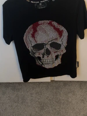 Svart t-shirt med dödskalletryck - Säljer en svart t-shirt med ett stort, detaljerat dödskalletryck i silver, rött och svart på framsidan. T-shirten har korta ärmar och en klassisk passform. Perfekt för dig som gillar edgy och unik stil.