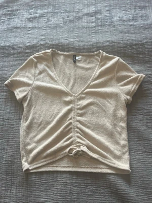 Beige croppad topp med snörning - Trendig beige croppad topp från H&M med v-ringning och snörning framtill. Tillverkad i mjukt material och har korta ärmar. Perfekt för dig som gillar en avslappnad men ändå snygg stil.