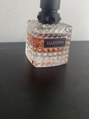 Valentino parfym med nitar och rosa ton - Säljer en lyxig parfym från Valentino i en fyrkantig glasflaska med nitar och struktur. Flaskan har en rosa tonad vätska och svart kork med nitar. Märket Valentino syns tydligt på etiketten framtill.