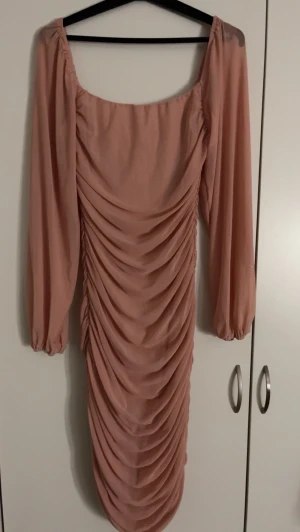 Ljusrosa långklänning med rynk - Trendig ljusrosa långklänning med rynkade detaljer framtill och långa, transparenta ärmar. Klänningen har en fyrkantig urringning och är tillverkad i ett lätt, flowy material som ger en elegant siluett. Perfekt för bröllop/fest 