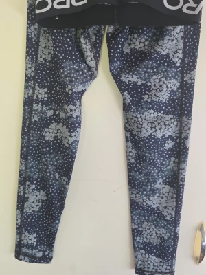 Mönstrade leggings från Nike - Snygga leggings från Nike med blå bas och vitt blommigt mönster. De har en bred resår i midjan med Nike-logga och sitter tight hela vägen ner. Perfekta för träning eller chill. Materialet är stretchigt och följsamt.