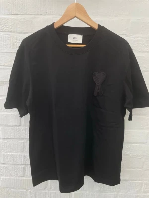 Svart t-shirt från Ami Paris  - Snygg svart t-shirt från Ami med deras ikoniska hjärtlogga broderad på bröstet. Tröjan är i nyskick med lappen på har inga tecken på slitage som fläckar eller hål. 