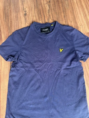 Marinblå t-shirt från Lyle & Scott - Snygg marinblå t-shirt från Lyle & Scott med klassisk gul logga på bröstet. T-shirten har rund halsringning och korta ärmar. Tillverkad i mjuk bomull som känns skön mot huden. Perfekt till jeans eller shorts för en clean look.