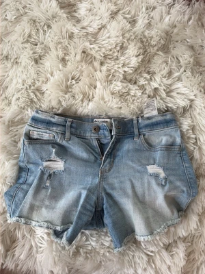 Ljusblå jeansshorts från Abercrombie & Fitch - Säljer ett par ljusblå jeansshorts med slitna detaljer och fransig kant. Shortsen är i mycket bra skick då de inte använts mycket. Storleken är 15/16 år men passar likadant som XS/S. Bara att höra av er om ni har frågor!!