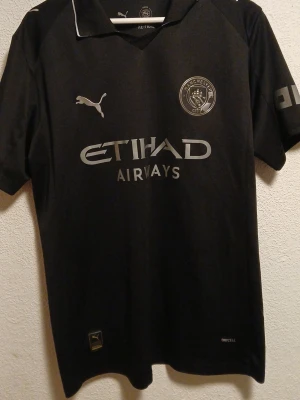 Svart Manchester City tröja Puma - Snygg svart Manchester City matchtröja från Puma med Etihad Airways-tryck på bröstet. Tröjan har korta ärmar, klubbmärke och Puma-logga framtill samt detaljer på ärmarna. Tillverkad i ett lätt och funktionellt material med Drycell-teknologi.