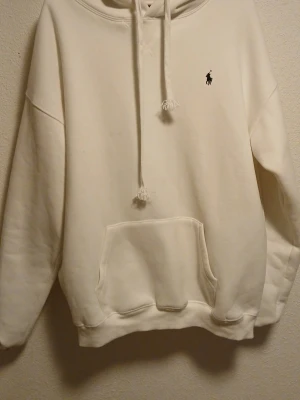 Vit hoodie från Polo Ralph Lauren - Snygg vit hoodie från Polo Ralph Lauren med klassisk broderad logga på bröstet. Hoodien har huva med dragsko och en stor magficka framtill. Tillverkad i mjukt bomullsmaterial som är skönt mot huden. Perfekt för en avslappnad och stilren look.