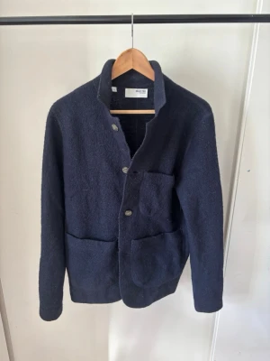 Mörkblå ull-overshirt med fickor - Stilren mörkblå overshirt i ullmaterial med tre stora utanpåliggande fickor och knäppning framtill. Rak passform och ståkrage ger en modern look. Perfekt lager på lager-plagg för kyligare dagar.