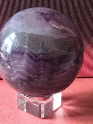 Fluorit klot med glas ställ - Säljer en rund Fluorit kula iolika nyanser av lila och vitt. Kulan har ett snyggt mönster och kommer med ett fyrkantigt ställ i genomskinligt glas eller akryl. Perfekt som dekoration eller för kristallintresserade.naturliga sprickor kan förekomma. Vikt 316 g, höjd 5 cm