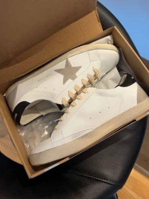 Golden Goose Superstar sneakers vit/beige - Golden Goose Superstar sneakers i vitt skinn med beige stjärna på sidan och beige snörning. Svart detalj på hälen och beige platt sula. Klassisk rund tå och GGDB-logga på plösen. Snygg och tidlös modell som passar till många stilar.