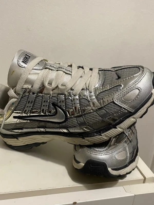 Nike p-6000 först till kvarl 550-600 - Snygga sneakers från Nike i metallic silver med svarta detaljer och vit sula. Skorna har ett futuristiskt mönster, meshpaneler och klassisk Nike Swoosh på sidan. Tjocka vita skosnören och vadderad plös för extra komfort. Perfekta för dig som gillar streetwear.