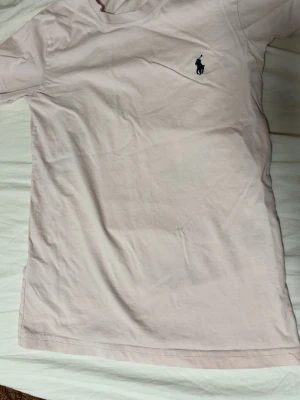 Ljusrosa t-shirt med broderad logga - Säljer en ljusrosa t-shirt med klassisk broderad logga på bröstet. T-shirten har rund halsringning och korta ärmar. Tillverkad i mjuk bomull, perfekt för en avslappnad och stilren look.