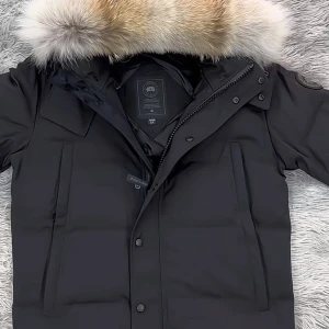 Svart dunjacka med pälskantad huva - Snygg svart dunjacka med stor huva som har en fluffig pälskant i beige och grå toner. Jackan har raka fickor framtill, dragkedja och knappar samt en stilren design. Perfekt för kalla vinterdagar och riktigt varm tack vare dunfyllningen. Skicka ett dm ifall du vill sänka priset 