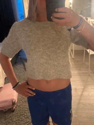 Grå cropped stickad tröja - Mysig grå stickad tröja med kort ärm och croppad passform. Tröjan har en enkel rund halsringning och är gjord i ett mjukt, fluffigt material som känns skönt mot huden. Perfekt för en avslappnad och trendig look.