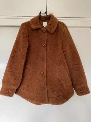 Brun teddyjacka från H&M - Säljer en brun teddyjacka från H&M i storlek 34. Jackan har klassisk krage, knäppning med bruna knappar och två fickor framtill. Materialet är mjukt och fluffigt, perfekt för kyliga dagar. Enkel och stilren design som är lätt att matcha.