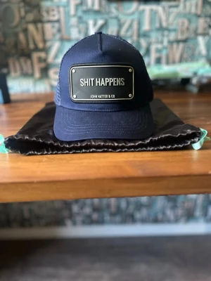 Oanvänd Marinblå keps John Hatter & Co - Snygg marinblå keps från John Hatter & Co med  mesh på sidorna och bak. Det är en rubber edition med texten 'SHIT HAPPENS'. Justerbar passform och klassisk trucker-keps.