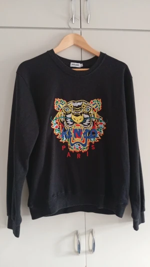 Kenzo sweatshirt  - Svart sweatshirt från Kenzo med ikoniskt broderat tigerhuvud i flera färger på bröstet och texten 'Paris' under. Rund halsringning, långa ärmar och mjukt material. Perfekt för dig som vill sticka ut med en färgstark detalj i din outfit.