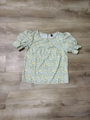 Blommig puffärm blus i pastellfärger - Söt blus med puffiga kortärmar och rund halsringning. Tyg i ljusgul och vit botten med småblommigt mönster i blått och gult. Perfekt för en fräsch och somrig look. Materialet känns lätt och luftigt.