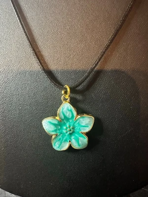 Halsband med turkos blomma - Unikt halsband med ett hängsmycke i form av en turkos blomma med guldfärgade kanter. Blomman har fem kronblad och små dekorativa detaljer i mitten. Hängs på ett svart rem som ger en stilren kontrast till det färgglada hänget. Perfekt för dig som gillar blommiga accessoarer.