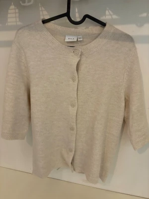 Beige kofta från Vila - Mysig beige kofta från Vila med rund halsringning och knappar framtill. Koftan har halvlånga ärmar och är tillverkad i ett mjukt, stickat material som känns skönt mot huden. Perfekt att slänga över en topp för en snygg outfit