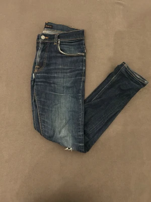 Thin finn - Snygga mörkblå nudie thin finn jeans med slim passform. Jeansen har litet hål mellan benen därefter det billiga priset. Strl 32/34 men passar som 30/32 kontakta gärna vid frågor eller funderingar