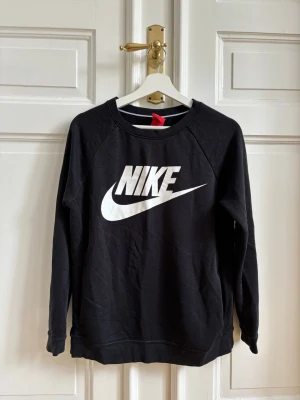 Svart Nike sweatshirt med logga - Svart sweatshirt från Nike med klassisk vit logga framtill. Tröjan har rund halsringning, långa ärmar, fickor och ribbade muddar vid ärmslut och nederkant. Tillverkad i mjukt bomullsmaterial för en avslappnad och sportig look.