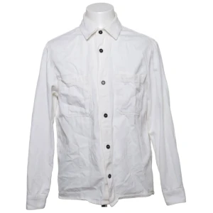 Vit overshirt med svarta knappar - Stilren vit overshirt med svarta knappar och två bröstfickor med lock. Klassisk krage och lång ärm, perfekt att bära öppen eller stängd. Overshirten har en rak passform och är tillverkad i ett lätt bomullstyg.
