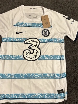 Chelsea FC bortatröja Nike vit/blå - Chelsea FC bortatröja från Nike i vitt med ljusblå och mörkblå horisontella ränder och klubbmärke på bröstet. Stort '3' sponsortryck på framsidan. Tillverkad i lätt och ventilerande material, perfekt för fotbollsträning eller match.