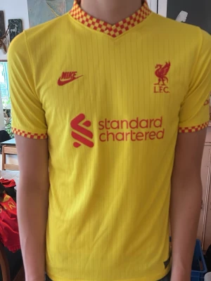 Liverpool FC gul matchtröja Nike - Gul Liverpool FC matchtröja från Nike med röda detaljer och röd-rutig krage och ärmslut. Tröjan har klubbmärke och Nike-logga på bröstet samt sponsortryck framtill. Tillverkad i lätt och ventilerande material, perfekt för fotboll eller träning. Modellen är 180 cm