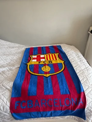 FC Barcelona handduk med klubbmärke - Säljer en färgstark handduk med FC Barcelonas klassiska klubbmärke och texten 'FC BARCELONA' längst ner. Handduken har blå och vinröda ränder och är tillverkad i mjukt tyg, perfekt för stranden eller gymmet. Köpt i spanien på FC Barcelonas butik.
