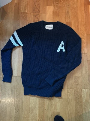 Marinblå stickad tröja Iconic Sport - Snygg marinblå stickad tröja från Iconic Sport med vita ränder på ena ärmen och ett stort A broderat på bröstet. Tröjan har rund halsringning och ribbade muddar. Perfekt för dig som gillar avslappnad stil.
