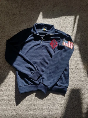 Jeffrey Epstein Halfzip 100% Bomull - Mörkblå stickad tröja med halv dragkedja, röd broderad logga på bröstet och amerikansk flagga på ärmen. Tröjan har ribbade muddar och är tillverkad i mjukt material. Perfekt för dig som gillar sportig och avslappnad stil.