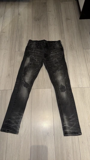 Svarta skinny jeans Supply & Demand - Säljer ett par svarta skinny jeans från Supply & Demand med slitningar och vita färgstänk för en edgy look. Jeansen har klassisk femficksmodell, knappgylf och är tillverkade i stretchigt denimtyg. Perfekta för dig som gillar streetwear och vill sticka ut.