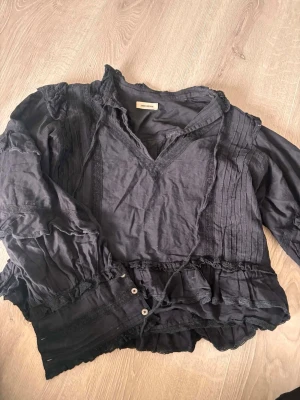 ZADIG & VOLTAIRE blus - Snygg svart blus från ZADIG & VOLTAIRE med volangdetaljer, spets och dekorativa sömmar. Blusen har v-ringning, långa ärmar med knappar vid ärmslut och plisserade partier framtill och baktill. Tillverkad i ett tunt, luftigt tyg som ger en avslappnad men chic look.